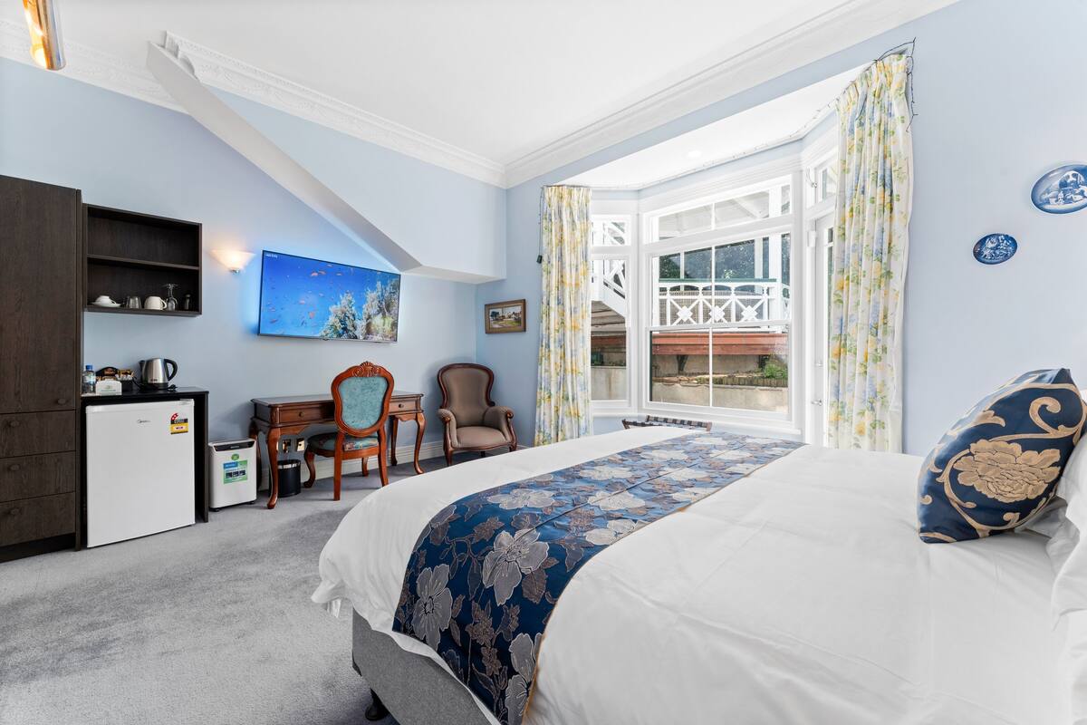 A bedroom with a bed, a desk, a chair, a small refrigerator, and a window with curtains.