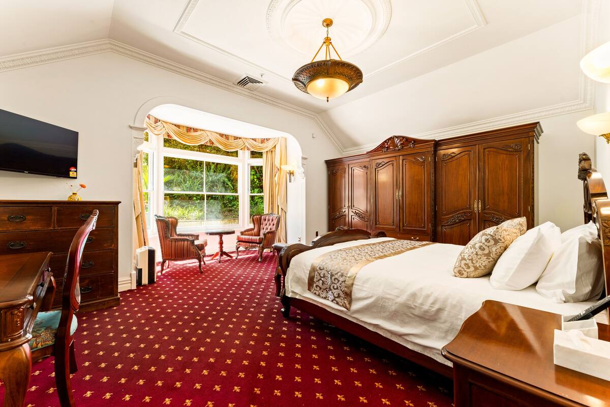 A hotel room with a large bed, a wooden wardrobe, a desk with a chair, a television, and a window with curtains.