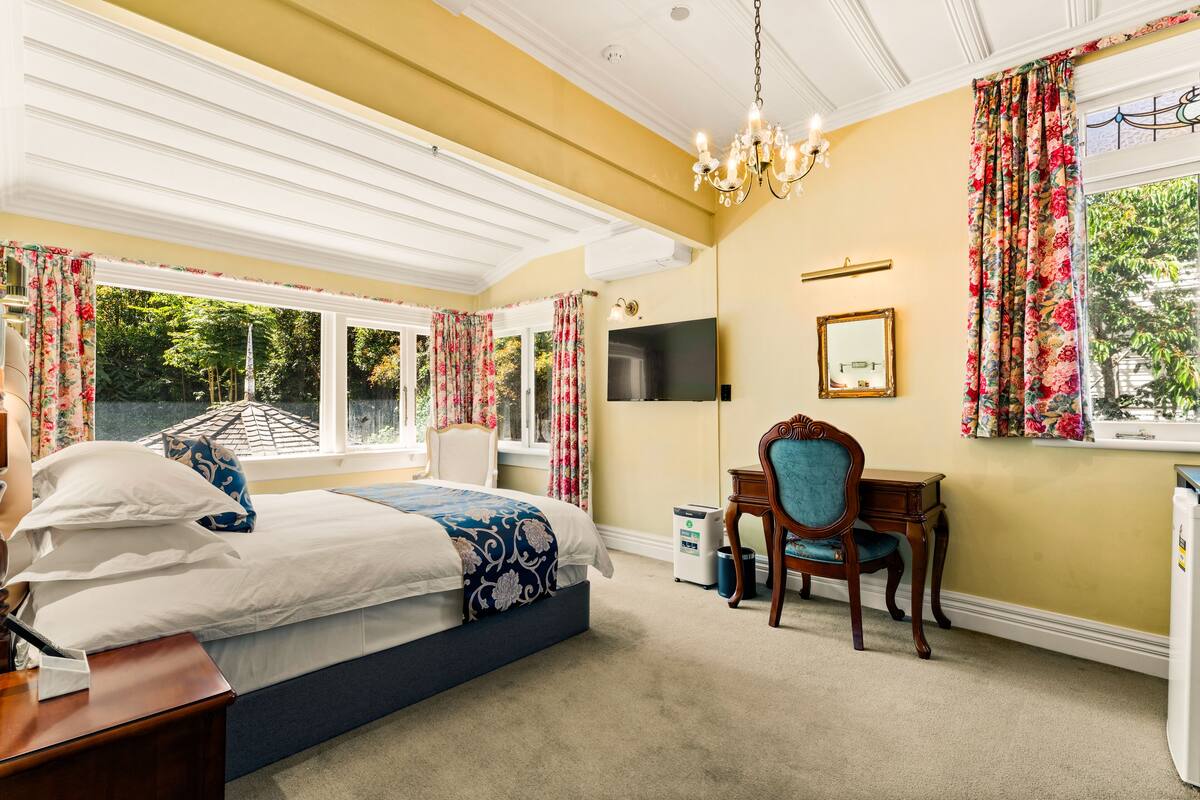 A bedroom with a bed, a desk, a chair, a TV, and a window with curtains.