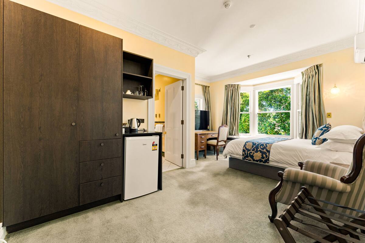A bedroom with a bed, a desk, a chair, a wardrobe, and a small refrigerator.