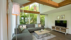 Ein modernes Wohnzimmer mit einem Ecksofa, einem Couchtisch, einem Fernseher auf einem Holzgestell und großen Fenstern mit Blick auf Grünflächen.