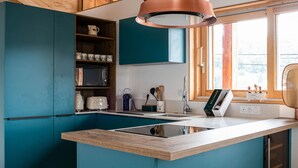 Una cucina moderna con mobili color petrolio, soffitto in legno, lampada a sospensione in rame e una finestra con vista.