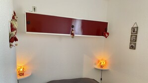 Une chambre à coucher avec un lit, deux lampes de chevet, une étagère rouge ornée d’objets décoratifs et une horloge murale.
