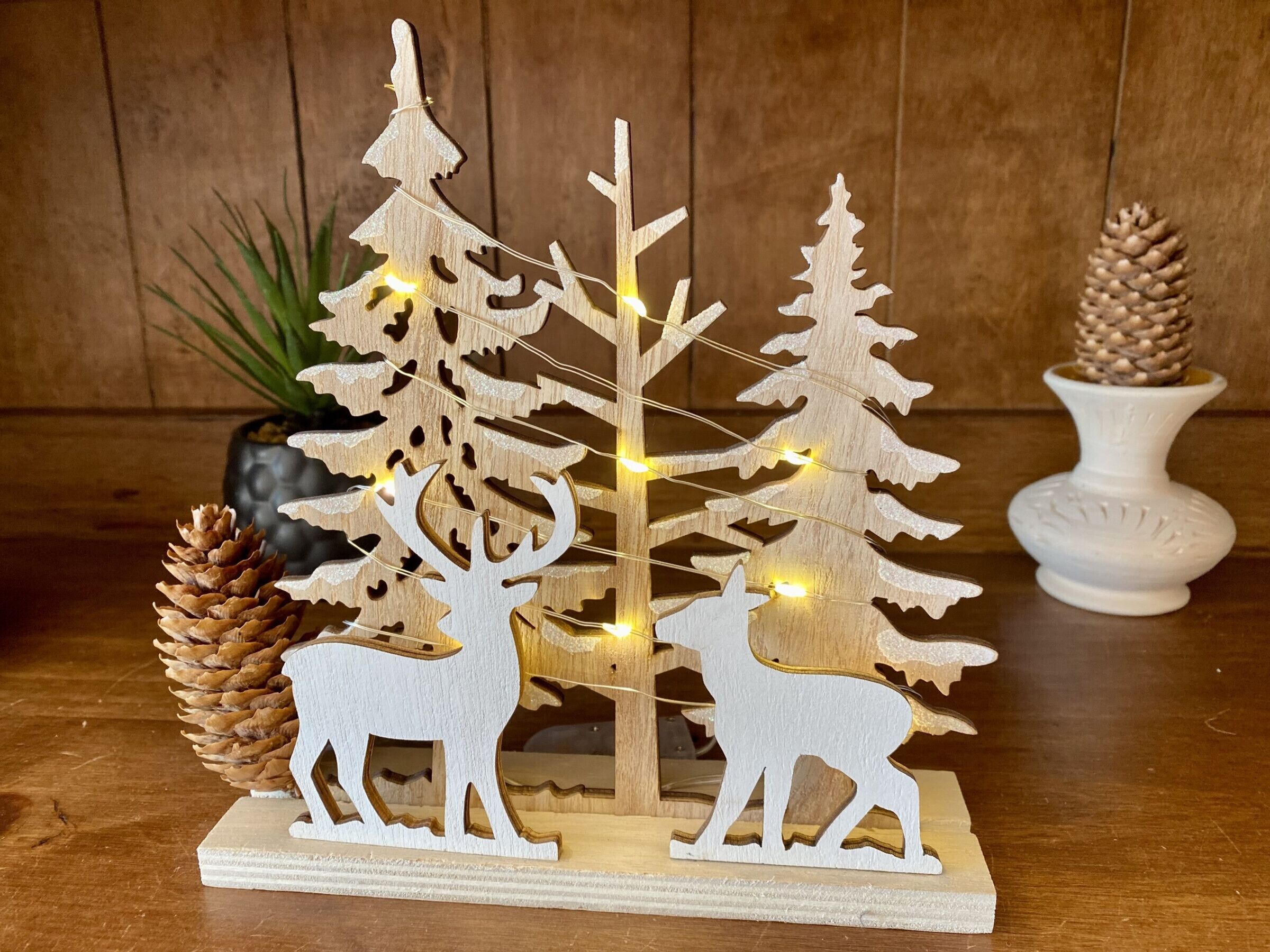 A wooden Christmas decoration with two deer figures, pine cones, and string lights.