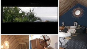Ein Zimmer mit schrägen Wänden, einem Bett mit gemusterten Textilien, einem runden Ventilator und einer Wanddekoration im Sonnenstrahl-Design.