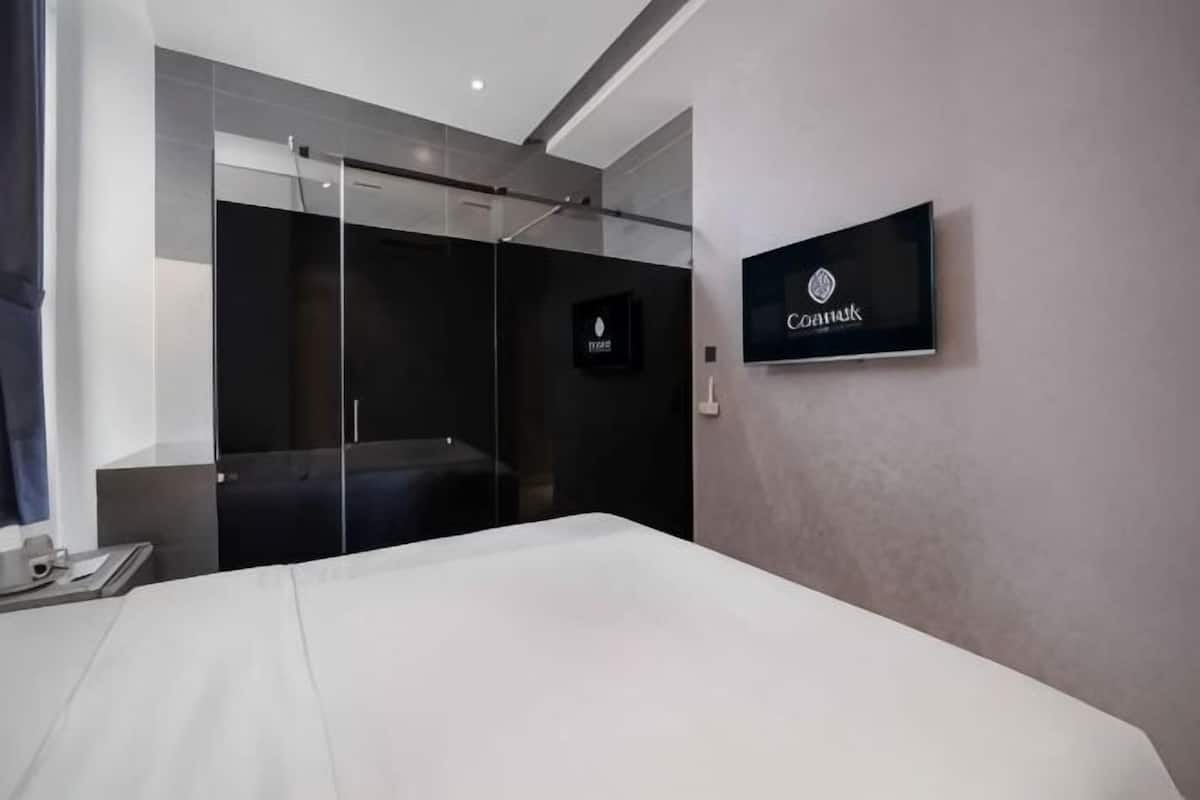 A modern hotel room with a bed, a glass shower enclosure, a mounted television, and a sign indicating the hotel name.