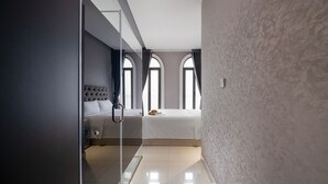 A modern bedroom with a glass partition, a tufted headboard, a large bed, and a view through arched windows.