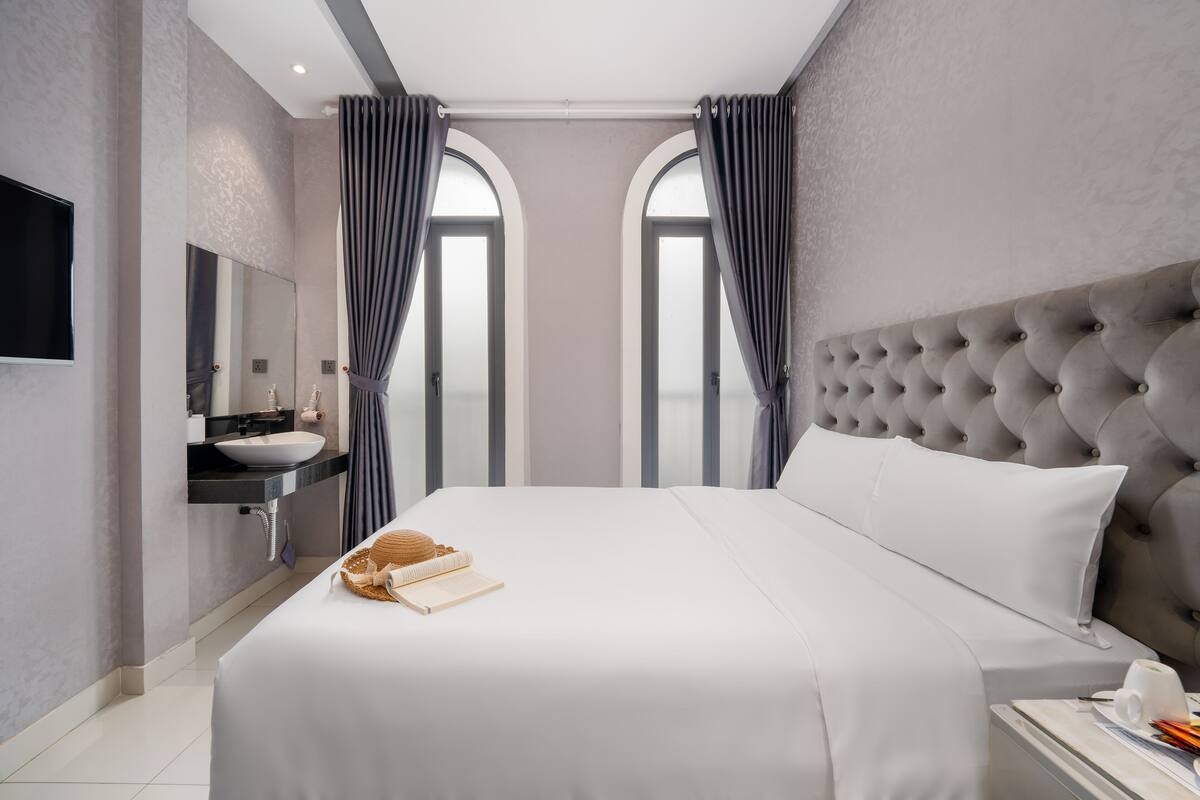 Une chambre d’hôtel moderne avec un grand lit, une coiffeuse avec un lavabo et deux fenêtres en arc surmont ées de rideaux.