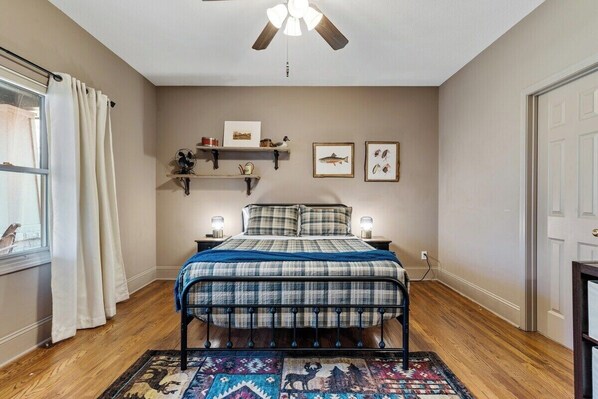 A bedroom with a bed, a ceiling fan, a window with curtains, wall-mounted shelves with pictures, and a rug.