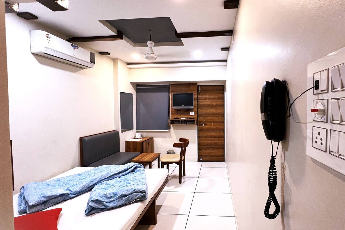 A compact hotel room with a bed, a small table, a chair, a desk, a TV, a wardrobe, and a wall-mounted phone.