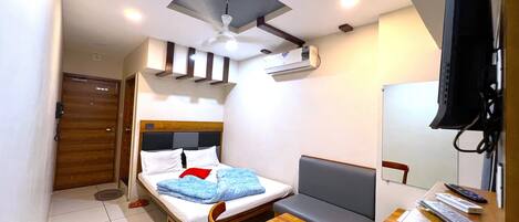 A compact hotel room with a bed, a small desk, a chair, a sofa, a ceiling fan, an air conditioning unit, and a television.