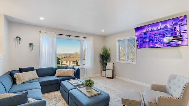 A modern living room with a sectional sofa, a small ottoman, a side table, and a chair. There is a large window with curtains, a potted plant, and a wall-mounted TV. - Draper Vacation Home - Families & Friends Welcome (Baraboo)