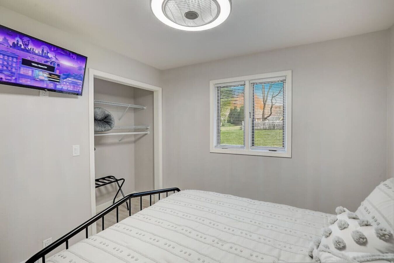 Ein Schlafzimmer mit einem Bett, einem an der Wand montierten Fernseher, einem Fenster mit Jalousien und einem Schrank mit Einlegeböden.