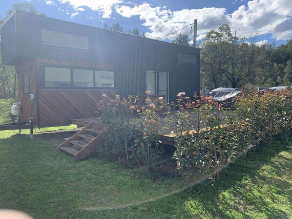 A modern two-story building with a flat roof, surrounded by a well-maintained garden with flowering plants and a wooden deck.