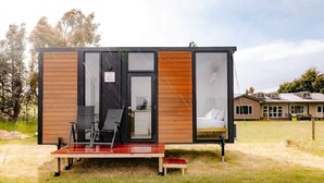 Eine moderne, kompakte Hütte mit Glastür, einer kleinen Sitzgelegenheit im Freien und einem sichtbaren Schlafzimmer mit Bett.