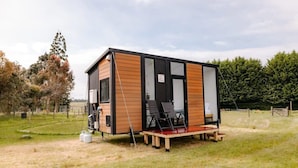 Eine kleine, moderne Holzhütte mit großen Fenstern, gelegen in einer grünen Wiese mit Bäumen im Hintergrund.
