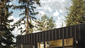 A modern building with large windows, a patio area with outdoor furniture, and a person tending to the garden. - Teakettle-Modern-Close To Glacier, Skiing (Columbia Falls)