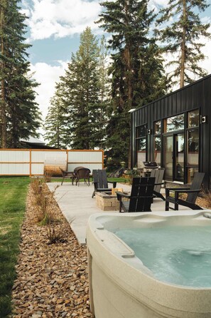 A modern outdoor patio with a hot tub, seating area, and a view of a forested area. - Teakettle-Modern-Close To Glacier, Skiing (Columbia Falls)