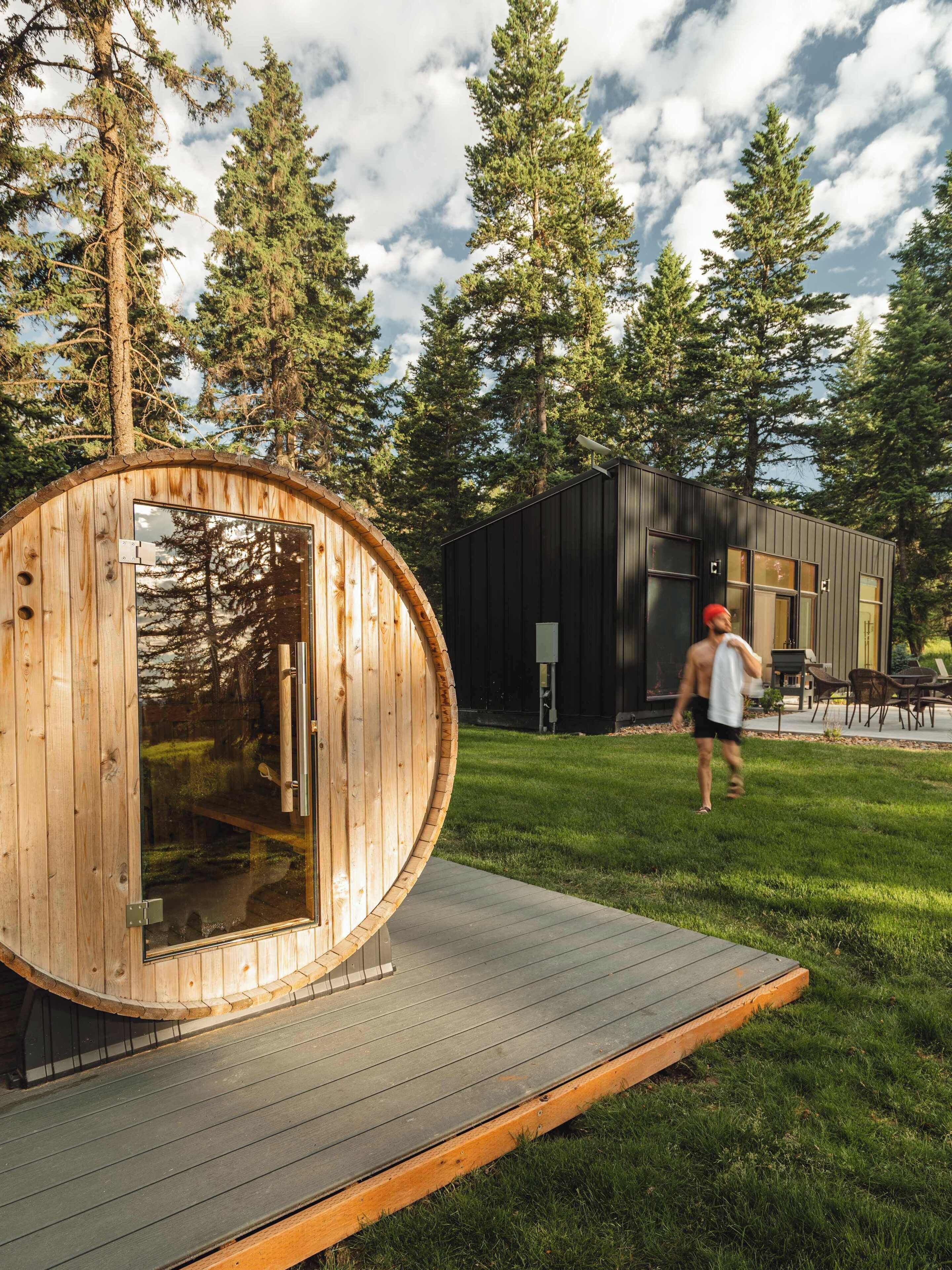 A wooden sauna structure with a glass door, a black building with windows, and a grassy area with a person walking.