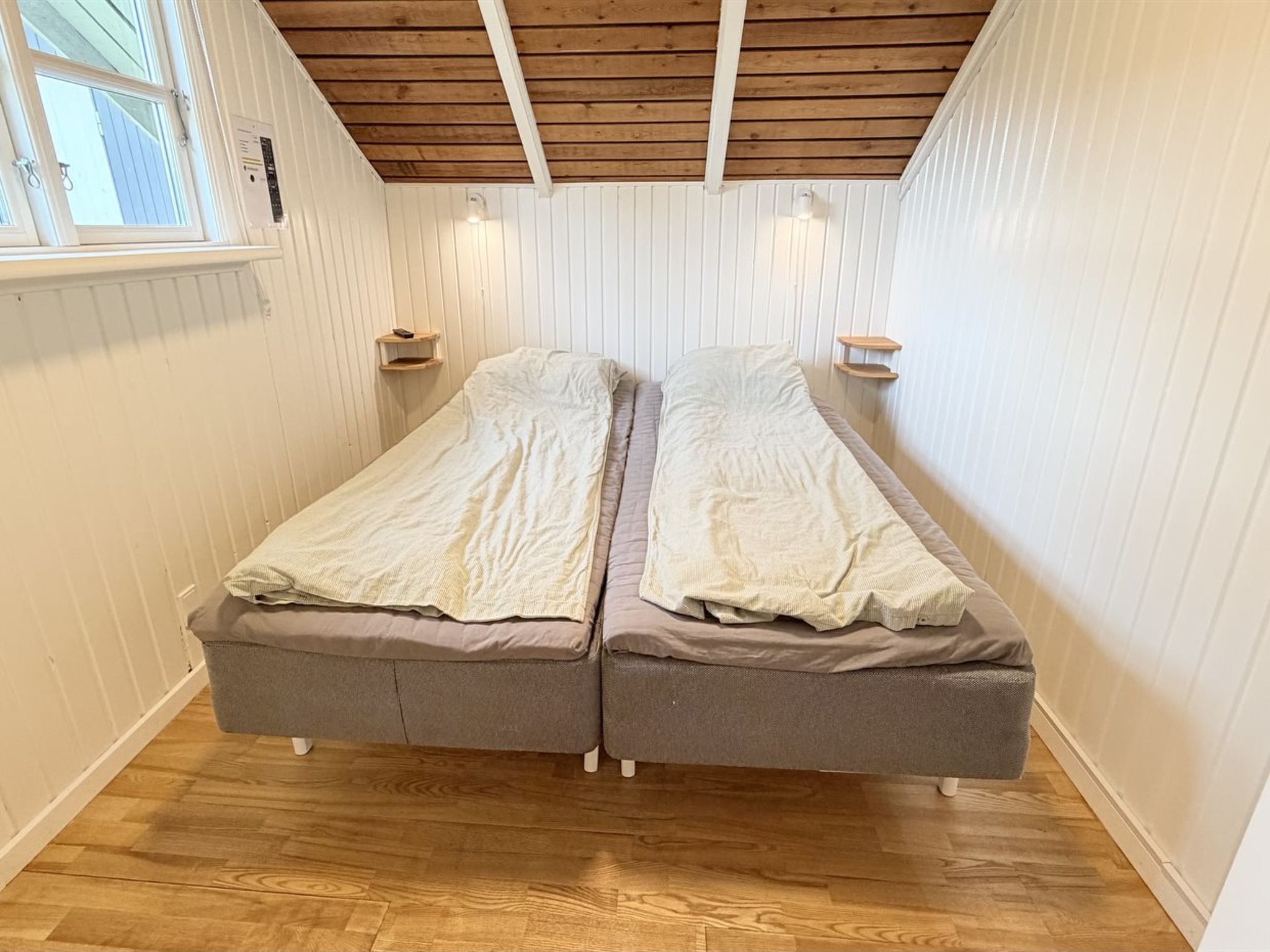 A small, single-room with two beds, wooden flooring, white walls, and a sloped ceiling.