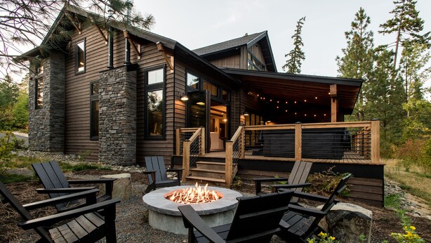 Una cabaña de troncos moderna con fachada de piedra, una terraza con barandilla de madera y un brasero rodeado de sillas Adirondack.