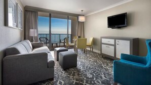A hotel room with a sofa, ottoman, a television, a desk, and a chair. - Seaside Splendor: 2nd Floor Oceanfront Queen Suite (Myrtle Beach)