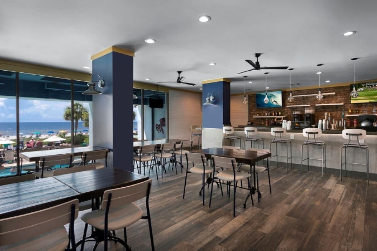Un restaurant moderne doté d’un plancher en bois, d’un bar et de grandes fenêtres offrant une vue sur la plage.