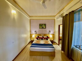 Una habitación de hotel con cama, dos mesitas de noche, ventilador de techo, un cuadro en la pared y una ventana con cortinas.