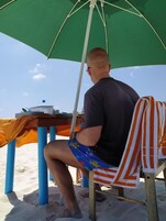 Una persona sentada bajo una sombrilla en la playa, con una mesa y una botella al frente.