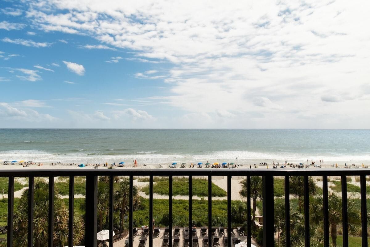 A beach view from a balcony with a railing, palm trees, and beach umbrellas.