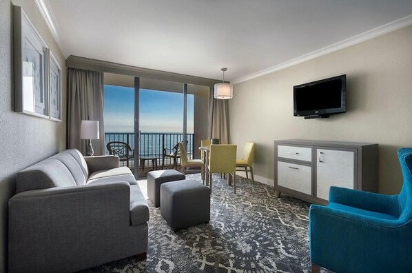 Une chambre d’hôtel avec un canapé, un pouf, une petite table, des chaises, une télévision et une vue sur l’océan.