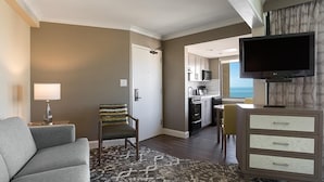 Habitación de hotel con sofá gris, alfombra estampada, sillón con cojín a rayas, televisor, minibar y vista al océano.