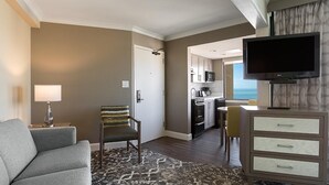 A hotel room with a gray sofa, a patterned rug, a chair with a striped cushion, a TV, a minibar, and a view of the ocean.