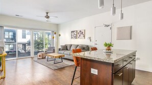 A modern kitchen with a central island, a dining area with a wooden table and chairs, and a living area with a sofa and a large window. - Stunning 1 Bedroom Luxury unit with a Pool (Columbus)