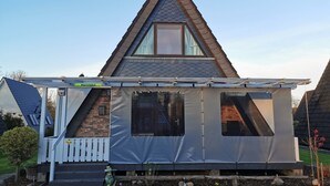 A house with a triangular roof, a covered porch, and a small garden area. - Modernes Nurdachhaus in Fedderwardersiel für bis zu 5 Personen und Hund (Butjadingen)