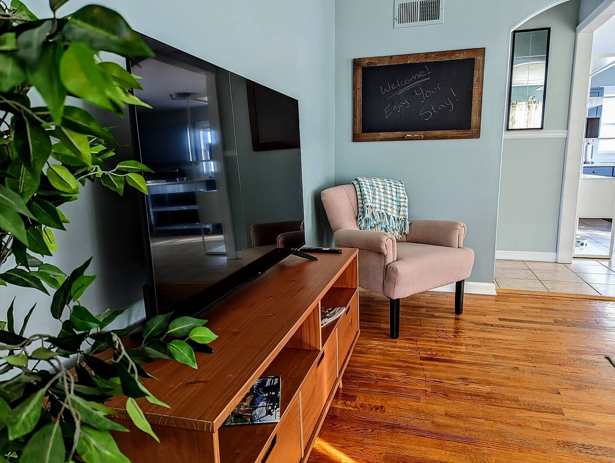 A living room with a flat-screen TV, a wooden entertainment unit, a pink armchair, and a chalkboard with a welcome message.