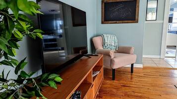 A living room with a flat-screen TV, a wooden entertainment unit, a pink armchair, and a chalkboard with a welcome message.