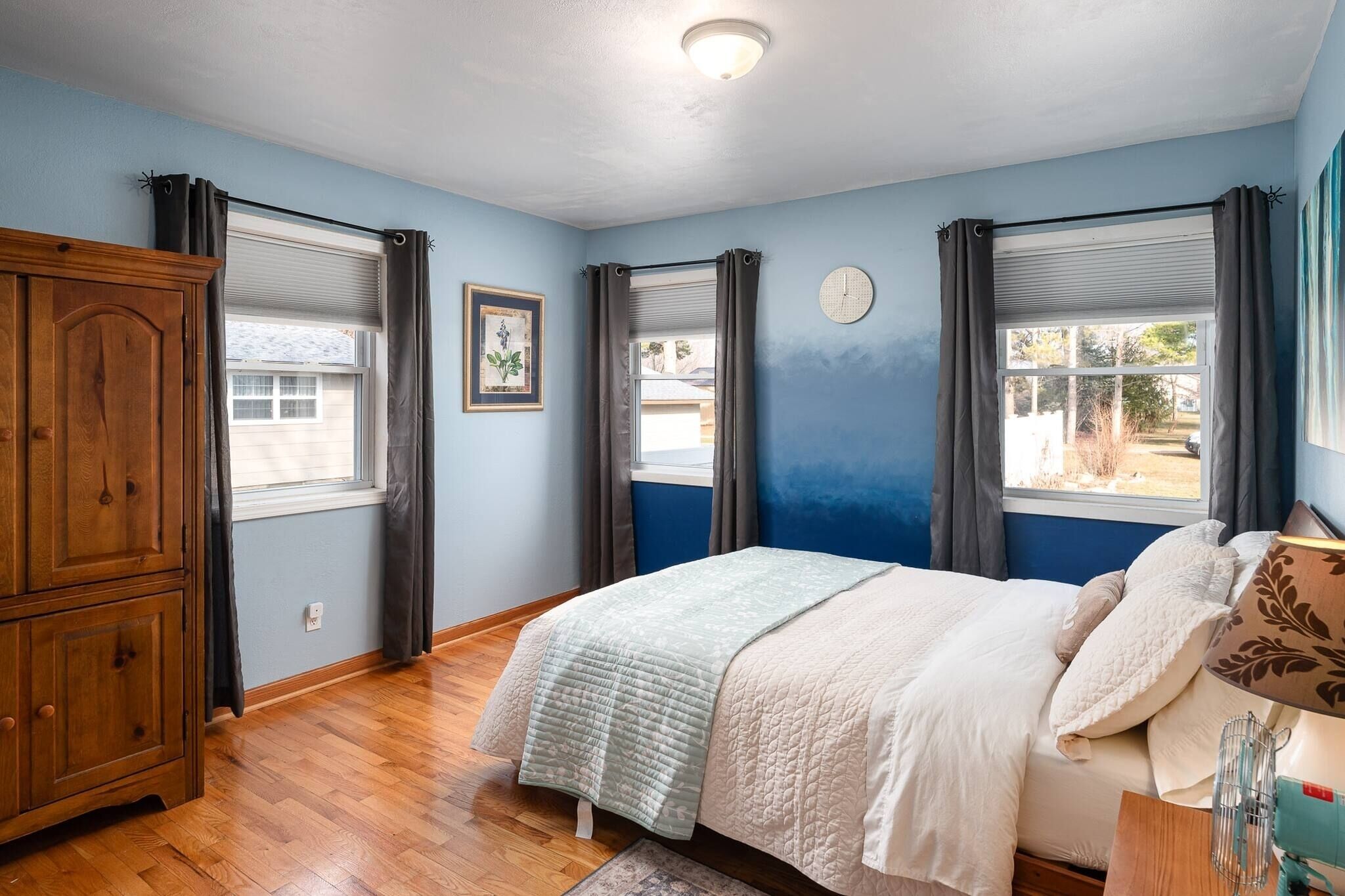 Un dormitorio con cama, armario de madera y dos ventanas con cortinas.