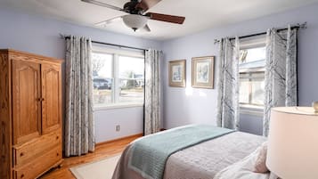 Un dormitorio con cama, ventilador de techo, armario de madera y dos ventanas con cortinas.