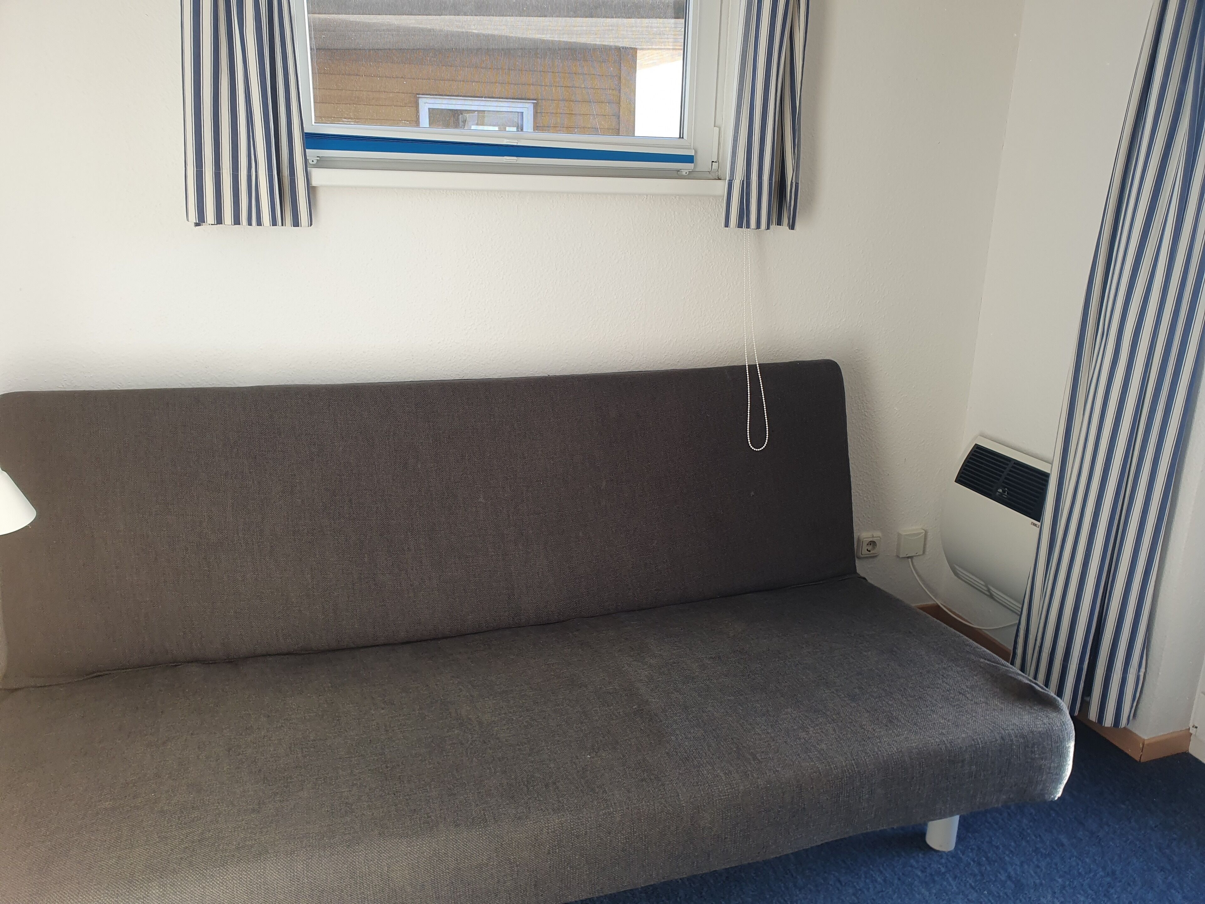 Ein Zimmer mit grauem Sofa, einem Fenster mit gestreiften Vorhängen, einer Wand-Klimaanlage und einem blauen Teppich.