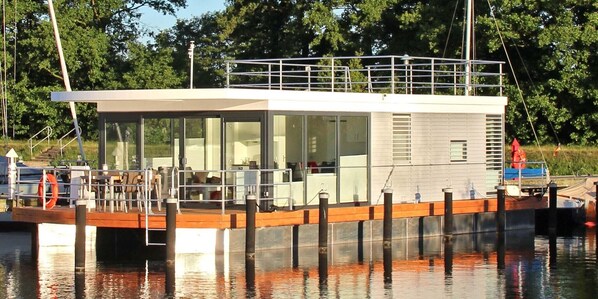 Ein modernes Hausboot mit Deck, großen Fenstern und einem überdachten Bereich.