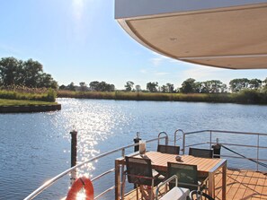 Ein Bootsdeck mit Tisch und Stühlen, einem Rettungsring und Blick auf einen ruhigen Fluss mit Bäumen und blauem Himmel.