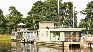 Ein Hausboot an einem Steg mit überdachter Terrasse und kleinem Balkon.
