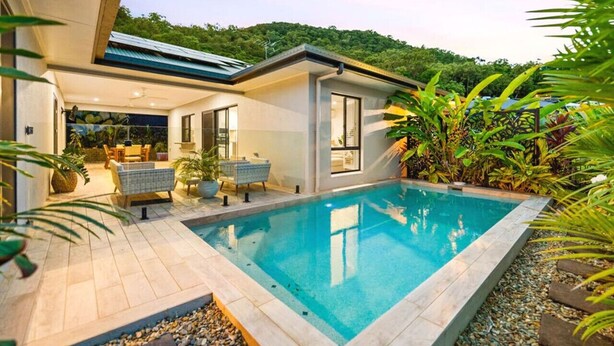 Ein modernes Haus mit Swimmingpool, umgeben von üppiger Vegetation und Blick auf einen bewaldeten Hügel.