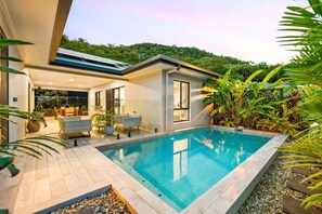 Une maison moderne dotée d’une piscine, entourée d’une végétation luxuriante et offrant une vue sur une colline boisée.