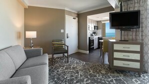 A hotel room with a gray sofa, a patterned rug, a TV on a stand, a chair, and a kitchen area visible through an open door. - Accessible Comfort with Ocean Views 6th Floor (Myrtle Beach)