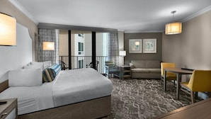 Ein Hotelzimmer mit Bett, Schreibtisch, Sofa und Esstisch. Vorhänge, Blick nach draußen und ein gemusterter Teppich sind ebenfalls vorhanden.