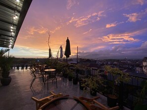 Terraço no telhado com área de estar externa, plantas em vasos e vista para o horizonte da cidade ao pôr do sol.
