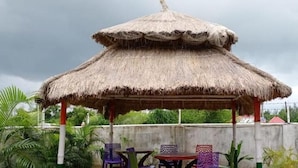 A thatched-roof gazebo with a table and chairs in a garden setting. - Swastik Resort (Kanthi)