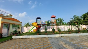 A playground with slides and swings, surrounded by potted plants and a building with a door and windows. - Swastik Resort (Kanthi)